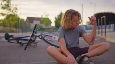 The boy sits on the road after falling off a bicycle, calms the pain in his elbow. Broken bike in the background. High quality 4k footage