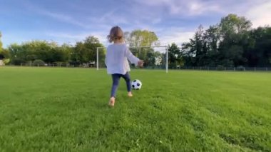 Küçük çocuk gün batımında futbol oynuyor, bebek yeşil futbol sahasında koşuyor ve topa vuruyor, kız çayırda koşuyor ve gülümsüyor, topa vurmayı öğreniyor, takım futbolcusu olmayı hayal ediyor..