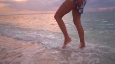 Sahilde yürüyen kadın bacakları, taze okyanus dalgalarının tadını çıkarıyor, büyüleyici pembe günbatımı yansıması. 