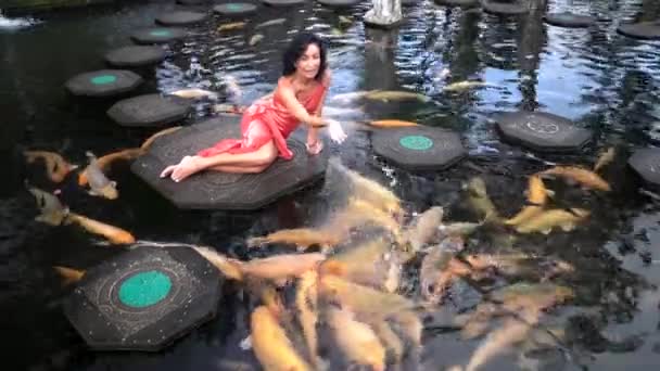 Элегантная счастливая женщина, лежащая на больших камнях в пруду, кормящая красочных рыб в воде, экзотический сад — стоковое видео