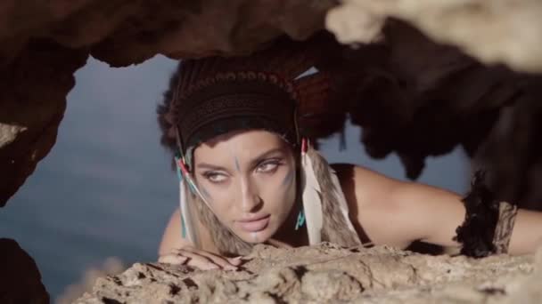 岩や狩猟に隠れて部族の装飾化粧や羽の帽子を持つ強力な無料の野生の女性 — ストック動画
