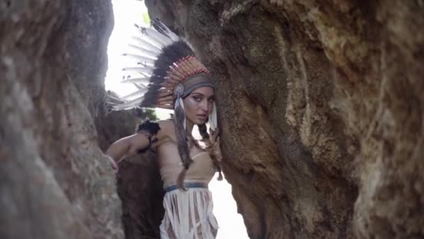 美しいモデルの女性が隠れて、岩の中にポーズをとって、羽の頭とカラフルな化粧をしています — ストック動画