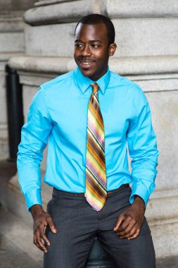 Açık mavi bir gömlek, renkli bir kravat, genç bir siyah işadamı gülümsüyor, mola vermek için dışarıda oturuyor.