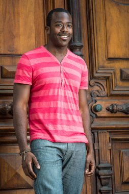 Kırmızı, pembe çizgili tişört, gri pantolon, bilezik takan, küçük bıyıklı genç siyah bir adam eski moda bir kapının önünde duruyor, gülümsüyor.