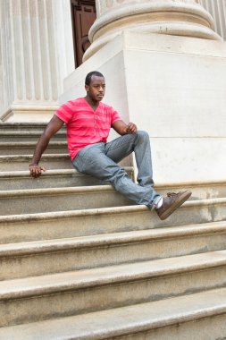 Kırmızı çizgili, pembe tişörtlü, gri pantolonlu, deri kahverengi ayakkabılı ve bilezik takan genç siyahi bir adam basamaklarda oturuyor ve şöyle düşünüyor: