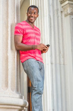 Kırmızı, pembe çizgili Henley V Neck tişörtlü, gri pantolonlu, bilezik takan genç bir siyah adam bir sütunun yanında duruyor, akıllı telefonundaki mesajları kontrol ediyor.