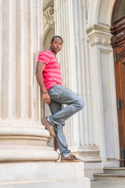 Kırmızı, pembe çizgili tişört, gri pantolon, deri ayakkabı giyen, bilezik takan, kampüsteki bir ofis binasının önünde kendinden emin bir şekilde bekleyen genç siyah bir öğrenci.