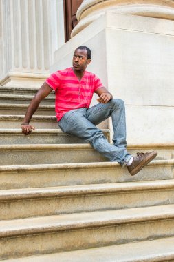 Bir bacağını esnetirken, genç siyah bir adam ofis binasının dışındaki merdivenlerde oturuyor, rahatlıyor ve düşünüyor.
