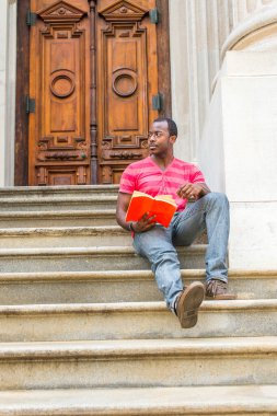 Bir bacağını uzatıyordu, elinde kırmızı bir kitap tutuyordu, genç siyah bir adam bir ofis binasının merdivenlerinde oturmuş, dinleniyor ve kitap okuyordu.