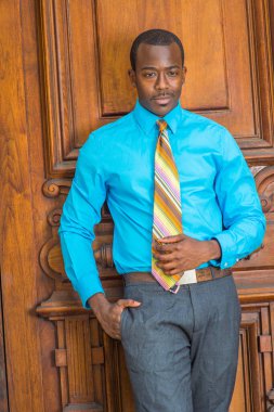 Açık mavi gömlek, gri pantolon, desenli kravat, cebine koyan bir el, siyah bir işadamı eski moda bir kapının yanında duruyor, aşağıya bakıyor, düşüncelerde kayboluyor.