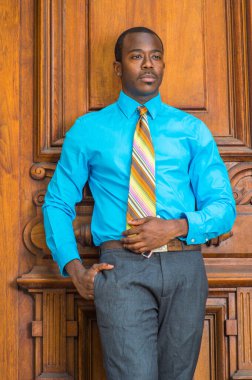 Açık mavi bir gömlek, gri bir pantolon, renkli bir kravat, cebine koyan bir el, genç siyah bir işadamı eski moda bir kapının yanında duruyor, düşüncelerde kaybolmuş.