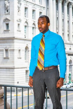 Açık mavi gömlekli, gri pantolonlu, renkli bir kravat takmış genç bir siyahi işadamı bir ofis binasının önünde, düşüncelere dalmış bir şekilde duruyor.