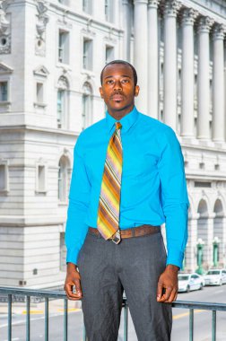 Açık mavi gömlekli, gri pantolonlu, renkli bir kravat takan genç bir siyahi işadamı bir ofis binasının önünde bekliyor.