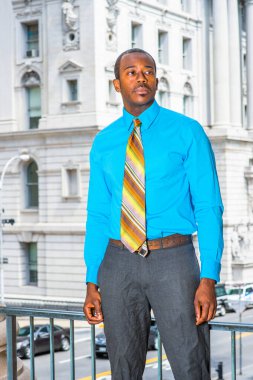 Açık mavi gömlekli, gri pantolonlu, renkli bir kravat takan genç bir siyahi işadamı bir ofis binasının önünde bekliyor.