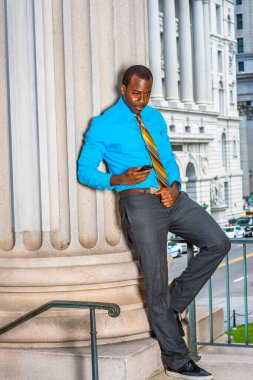 Açık mavi gömlekli, gri pantolonlu, deri ayakkabılı, desenli kravatlı genç bir iş adamı ofis binasının önünde durmuş cep telefonundaki mesajları kontrol ediyor.