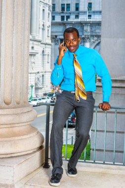 Açık mavi gömlekli, gri pantolonlu, deri ayakkabılı, desenli kravatlı genç siyahi bir işadamı ofis binasının dışında oturuyor ve cep telefonuyla konuşuyor.