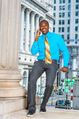Açık mavi gömlekli, gri pantolonlu, deri ayakkabılı, desenli kravatlı genç siyahi bir işadamı ofis binasının dışında oturuyor ve cep telefonuyla konuşuyor.
