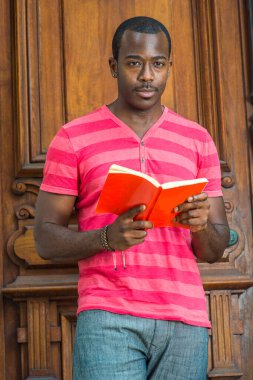 Kırmızı, pembe çizgili Henley V Neck tişörtü, gri pantolon, bilezik takan, küçük bıyıklı genç, yakışıklı bir siyahi adam eski moda bir kapının önünde kitap okuyor.