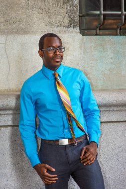 Açık mavi bir gömlek, renkli bir kravat, gri pantolon, gözlük takan genç bir siyahi üniversite öğrencisi kampüs duvarının önünde kendinden emin bir şekilde bekliyor.
