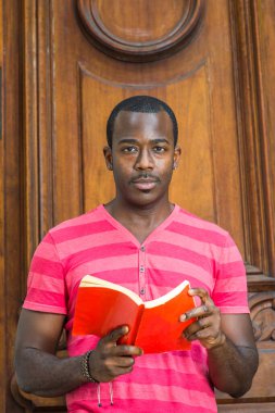 Kırmızı, pembe çizgili Henley V Neck tişörtü, gri pantolon, bilezik takan, küçük bıyıklı genç, yakışıklı bir siyahi adam eski moda bir kapının önünde kitap okuyor..