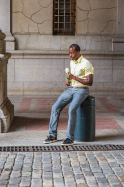 Kaşlarını çatmış, sabırla, açık sarı gömlekli, gri pantolonlu, siyah ayakkabılı, genç, çekici siyah bir adam, beyaz bir gül tutuyor, eski şehirde caddenin kenarında seni bekliyor.