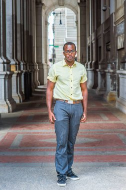Açık mavi gömlekli, desenli kravatlı, gri pantolonlu, gözlüklü siyahi bir üniversite öğrencisi kampüsün girişinde dikilip sana bakıyor.