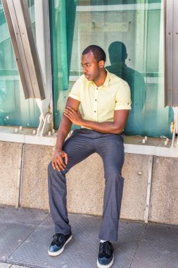 Açık sarı gömlekli, gri pantolonlu, spor ayakkabılı genç, çekici bir zenci metal bir yapının yanında oturuyor ve camdan bir duvara yaslanıyor.