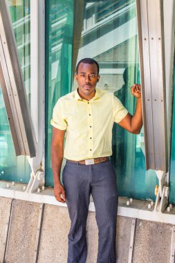 Açık sarı gömlekli, gri pantolonlu, genç, çekici siyah bir adam bir üçgen yapının yanında duruyor ve sana bakıyor.