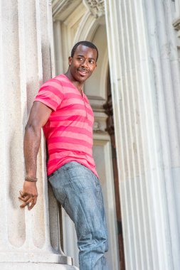 Kırmızı, pembe çizgili Henley V Neck tişörtlü, gri pantolonlu, bilezik takan genç siyah bir adam bir sütunun yanında duruyor, gülümsüyor ve seni bekliyor.