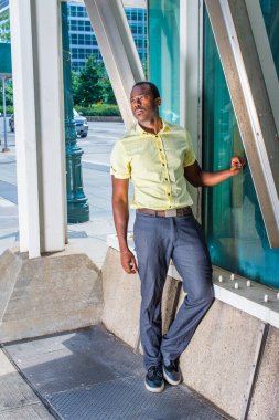 Açık sarı gömlekli, gri pantolonlu ve siyah ayakkabılı genç bir adam cam bir duvarın yanında dikiliyor ve sokağın köşesinde metal bir yapıya yaslanıyor.