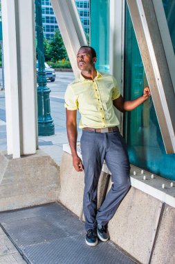 Açık sarı bir gömlek, gri pantolon ve siyah ayakkabılar giyen genç bir siyah adam sokağın köşesindeki metal bir yapıya karşı cam bir duvarın yanında duruyor ve derin düşüncelere dalıyor..