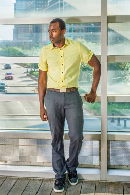 Açık sarı bir gömlek, gri pantolon ve siyah ayakkabılar giyen genç bir siyah adam derin düşüncelere dalmak için büyük bir cam duvarın yanında duruyor. Arka plan bir sokak sahnesinin üzerinde.