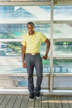 Açık sarı bir gömlek, gri pantolon ve siyah ayakkabılar giyen genç bir siyah adam sokağın köşesindeki metal bir yapıya karşı cam bir duvarın yanında duruyor ve derin düşüncelere dalıyor.