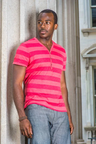 Dressing in red, pink stripe Henley V Neck T shirt,  gray pants, wearing a bracelet, a young black guy is standing by a pillar, confidently looking forward