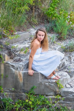 Beyaz mavi bir elbise, beyaz bir kemer ve sandalet giymiş, güzel sarışın bir kız vahşi, izole bir alanda oturuyor, derin düşüncelere dalıyor.