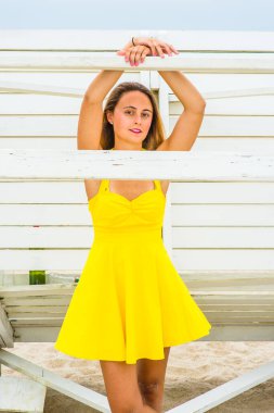 Parlak sarı bir elbise giyip, iki kolunu ve elini tahta bir sopaya koyan genç ve güzel bir kız sahilde beyaz bir yapının yanında duruyor ve sana bakıyor.