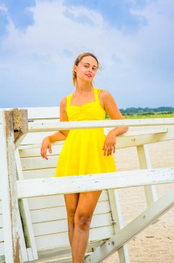 Parlak sarı bir elbise giymiş, iki eli saçlarını tarıyor, genç ve güzel bir kadın sahilde beyaz bir yapıya karşı duruyor, eğleniyor.