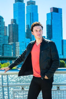 Siyah deri ceket, siyah kot pantolon ve açık kırmızı tişört giyen genç yakışıklı bir adam nehir kenarında dinleniyor ve düşünüyor. Arka planda büyük şehir yüksek binaları var..