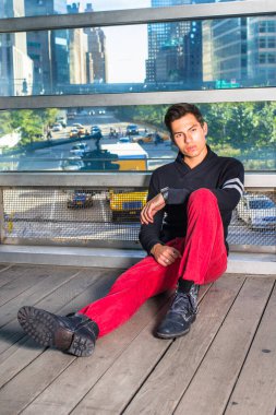 Siyah şal yakalı genç adam, kırmızı kot pantolon, siyah deri ayakkabılar, New York 'ta büyük cam duvarın yanında oturuyor, dinleniyor, düşünüyor. Arka plan işlek sokak manzarası.