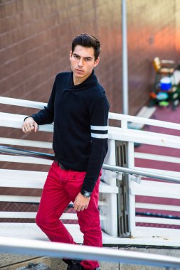 Siyah şal yakalı ve kırmızı kotlu genç bir adam beyaz bir çitin yanında duruyor, kendinden emin bir şekilde ileriye bakıyor.