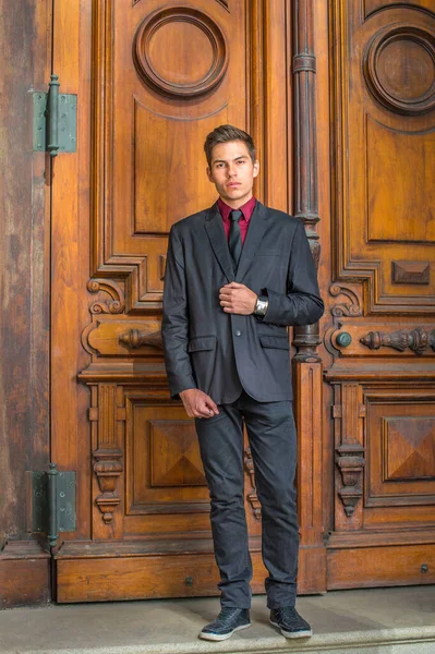 Dressing in a red undershirt, a black blazer, black jeans and a black tie, a young handsome college student is standing in the front of old fashion style door, looking forward