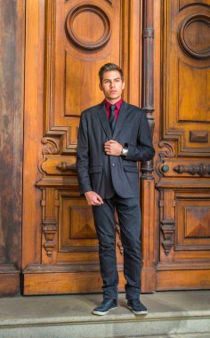 Kırmızı fanilalı, siyah ceketli, kot pantolonlu, kravatlı, deri ayakkabılı yakışıklı bir üniversite öğrencisi eski moda kapısının önünde duruyor.