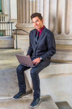 Kırmızı fanila, siyah ceket, kot pantolon, siyah kravat giyen genç bir üniversite öğrencisi kampüsteki bir ofisin dışında dizüstü bilgisayarla çalışıyor.