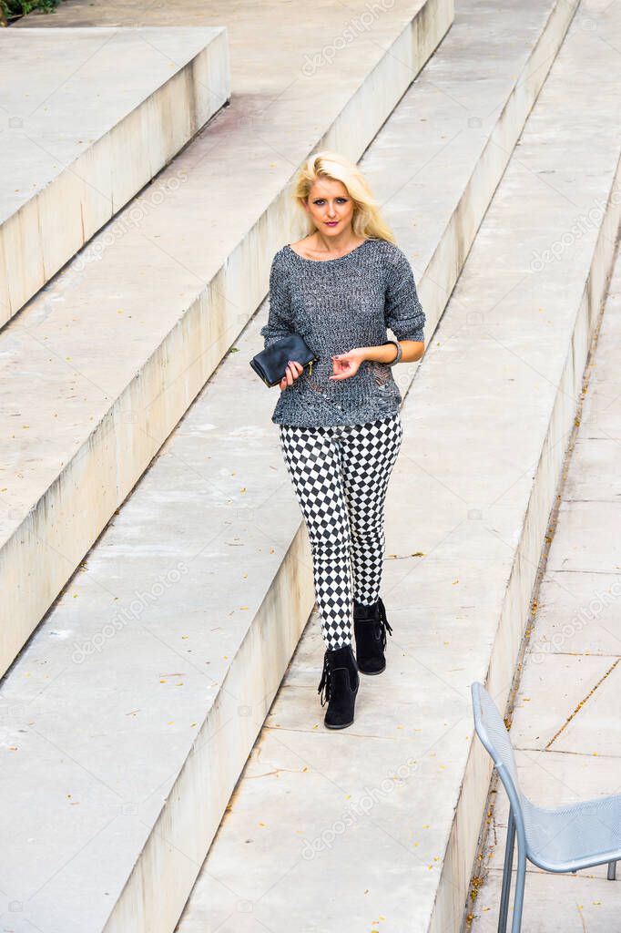 Dressing in a gray fashionable sweater, black and white pattern pants, black boots, holding a black leather purse, a young blonde girl is walking on steps