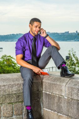 Mor gömlekli, gri pantolonlu, siyah kravatlı, elinde kırmızı bir kitap olan, sakallı ve bıyıklı bir üniversite öğrencisi kampüste bir nehrin kenarında oturmuş telefonla konuşuyor.