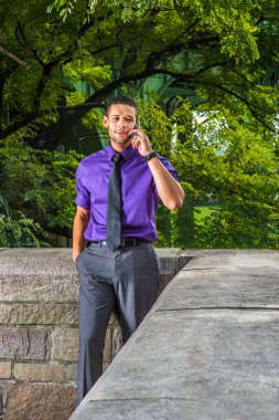 Mor gömlekli, gri pantolonlu, siyah kravatlı, sakallı, genç ve yakışıklı bir iş adamı dışarıda cep telefonuyla konuşuyor.