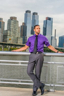 Mor gömlekli, gri pantolonlu, siyah kravatlı, deri ayakkabılı, sakallı ve bıyıklı genç bir iş adamı kalabalık bir iş bölgesinin dışında durup dinleniyor ve düşünüyor.