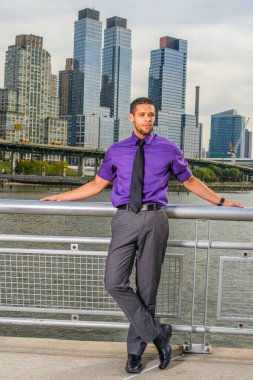 Mor gömlekli, gri pantolonlu, siyah kravatlı, deri ayakkabılı, sakallı ve bıyıklı genç bir iş adamı kalabalık bir iş bölgesinin dışında durup dinleniyor ve düşünüyor.