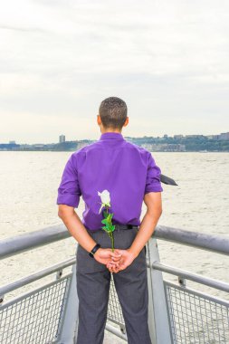 Sırtında çiçek tutan genç adam. Mor gömlekli, gri pantolonlu genç bir adam, nehrin kenarında duruyor, suya bakıyor, arkasında beyaz bir gül tutuyor, seni bekliyor.