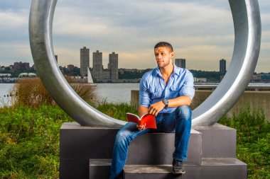 Dışarıda okuyan adam. Açık mavi gömlek, mavi kot pantolon, kırmızı kitap tutan eller, küçük sakallı ve bıyıklı genç bir üniversite öğrencisi nehir kenarında oturuyor, okuyor, kaşlarını çatıyor, düşünüyor.